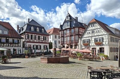 Photograph showing a historic European town square lined with colourful half-timbered buildings, cafes, and flower boxes