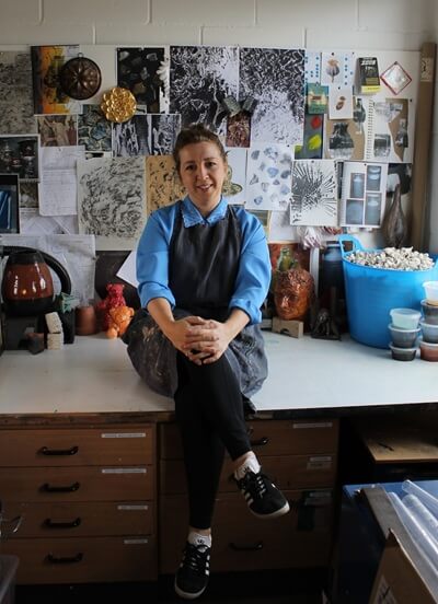 Catherine Bridget Phillips, dressed in a blue shirt and apron, sits cross-legged on a worktable in an art studio, surrounded by art materials, ceramics, and a wall covered with photos, sketches, and inspiration images.