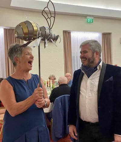 Photograph captures a smiling woman holding a large metal bee sculpture on a metal pole with a bearded man at a gala event
