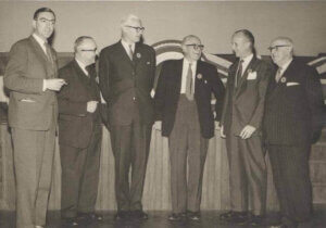 Black-and-white group photograph shows six formally dressed men standing on a stage at a civic event