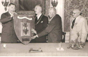 Black-and-white photograph shows four men at a formal presentation, holding a large decorative shield or plaque