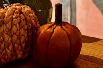 Two decorative pumpkins, one woven from straw-like material and the other made of soft orange fabric, sit on a wooden surface with a blurred background featuring warm colours—perfect festive touches for a cosy Halloween Pumpkin Trail for children.