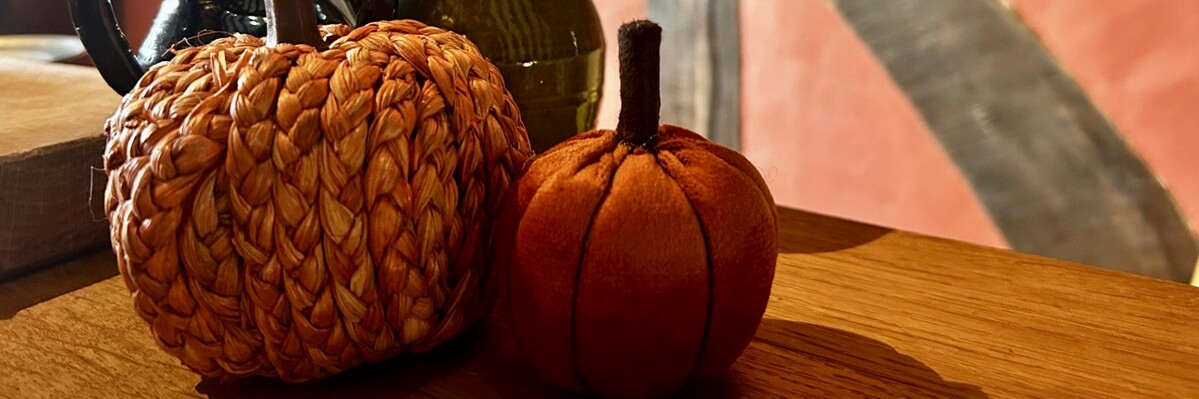 Two decorative pumpkins, one woven from straw-like material and the other made of soft orange fabric, sit on a wooden surface with a blurred background featuring warm colours—perfect festive touches for a cosy Halloween Pumpkin Trail for children.