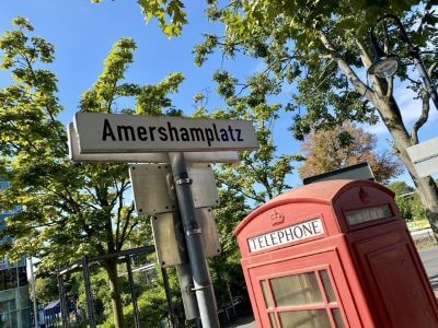 Photograph showing Amershamplatz street sign beside a red British telephone box with leafy trees and blue sky