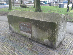 Photograph of a weathered rectangular stone monument with moss-covered surfaces and a bronze plaque set into its front