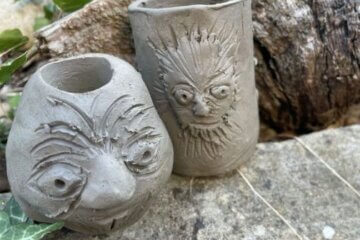 Two handmade grey clay vessels with carved faces on their surfaces.