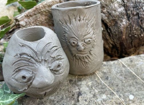 Two handmade grey clay vessels with carved faces on their surfaces.
